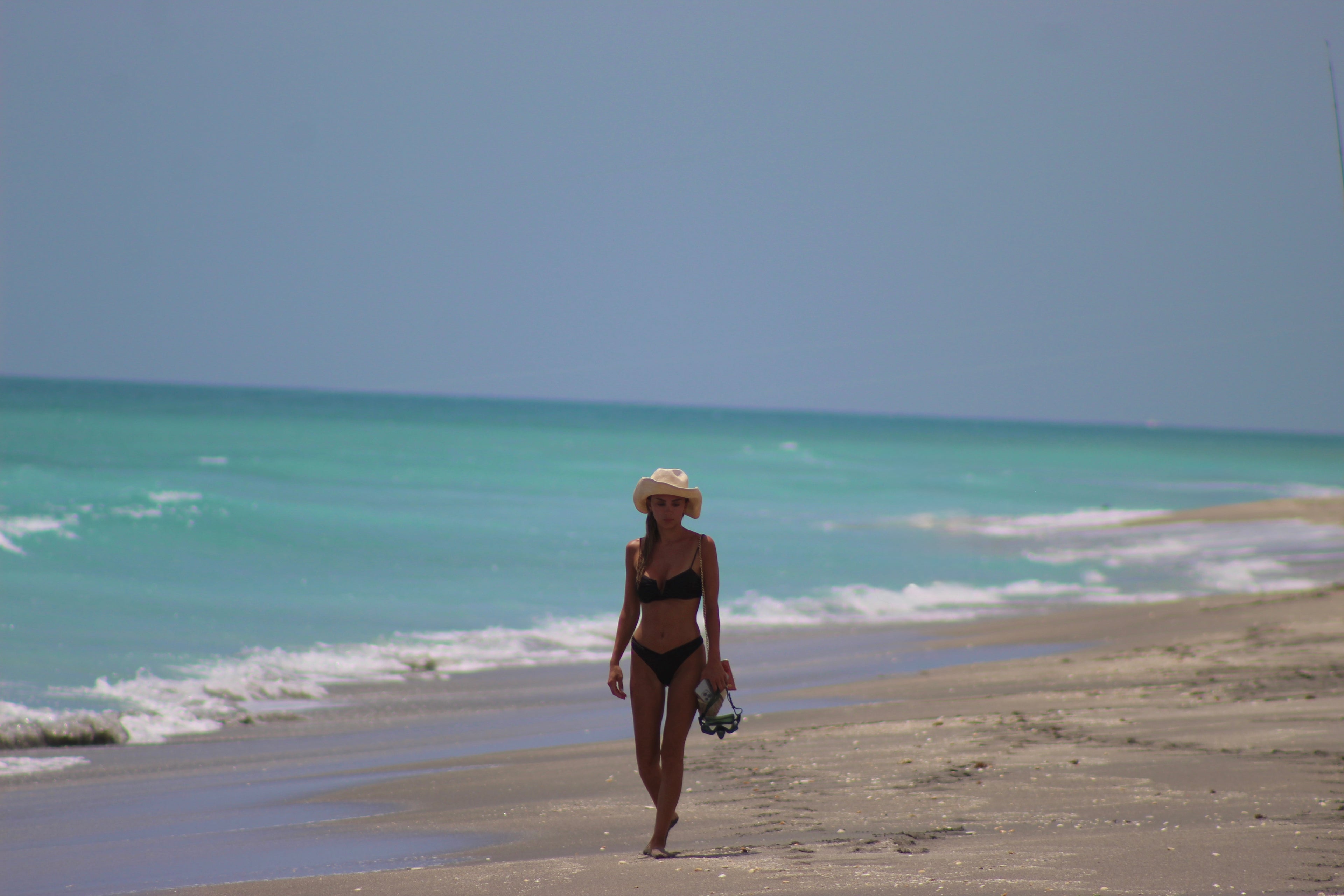 Woman in a bikini and hat walking on a beach with clear blue water and sky.